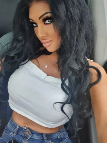 BritishTastyCherryxo with long wavy dark hair wearing a white tank top and blue jeans while sitting in a car.