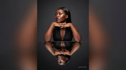 ZoeySpark poses with her hands under her chin, wearing a black low-cut top against a dark background with a clear reflection below.