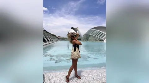 KaylaBryan poses in a black swimsuit and sun hat by a large outdoor pool and modern white buildings under a blue sky.