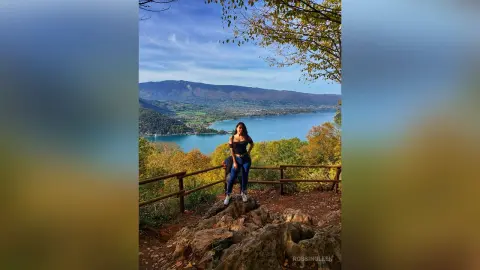 RossiNoleen stands by a wooden fence overlooking a blue lake and mountains, wearing a black tank top and blue jeans.