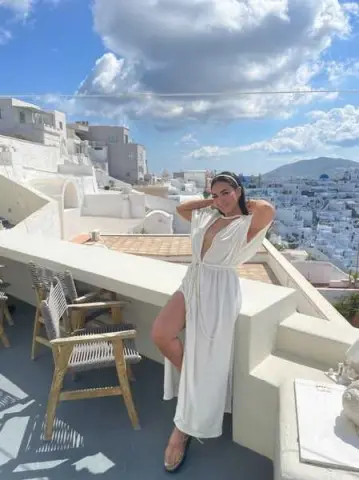 Stylish woman in a white maxi dress posing on a sunny Santorini balcony overlooking white Mediterranean buildings.