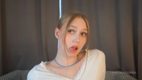 Blonde woman in a white lace top making a playful expression against a dark curtain background.