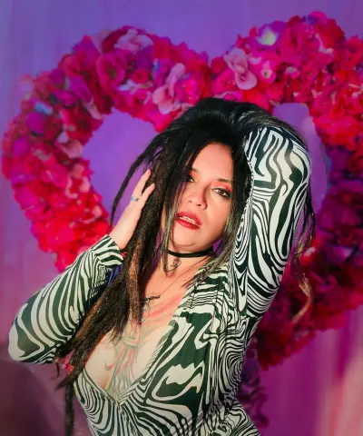 Woman with long dreadlocks in a black and white patterned dress posing against a pink floral heart backdrop.