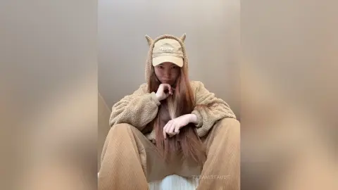 Young woman in cozy tan fleece loungewear and matching cap sitting relaxed in a minimalist indoor setting.