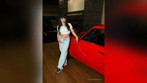 Stylish woman in white crop top and light jeans posing next to a red car in a modern garage.