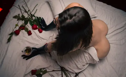 Top view of a woman in black gloves and white shirt lying on bed with red roses and a glass of wine.