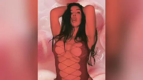 Glamorous woman in a sheer red lace bodysuit posing against a soft pink dreamy background.
