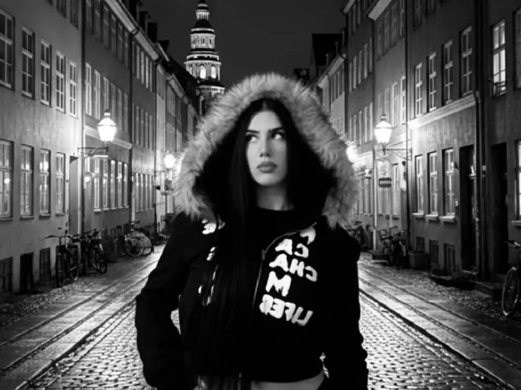 Moody black and white portrait of a woman in a fur-lined hoodie standing on a cobblestone city street at night.