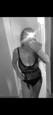 Woman in black fishnet bodysuit taking a moody black and white mirror selfie indoors.