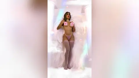 LinaVon poses naked with pink flower pasties and light pink thong, wearing sheer white stockings against a soft white background.