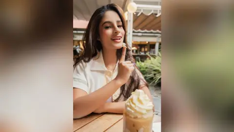 TefyRusso smiles while sitting at a table, touching her finger to her lips with long brown hair and a white collared shirt.