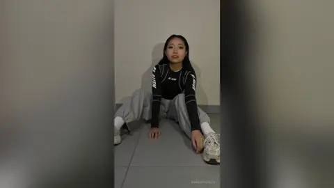 Young woman in a black long-sleeve graphic shirt and grey sweatpants sitting on a floor in a minimalist room.