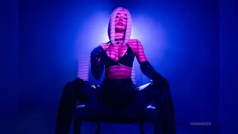 HanaRoss poses in a black leather bra and gloves under blue and pink neon lights.