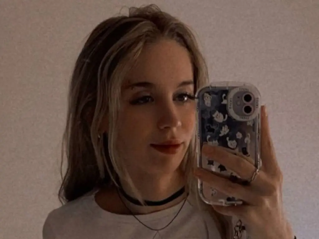 Young woman with blonde hair and black choker taking a moody selfie in a patterned phone case.