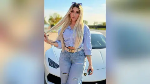 Blonde woman in striped shirt and denim jeans posing confidently by a white luxury sports car outdoors.