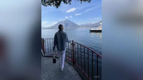 Woman in a denim jacket and white pants looking at the scenic lake and mountains from a balcony.