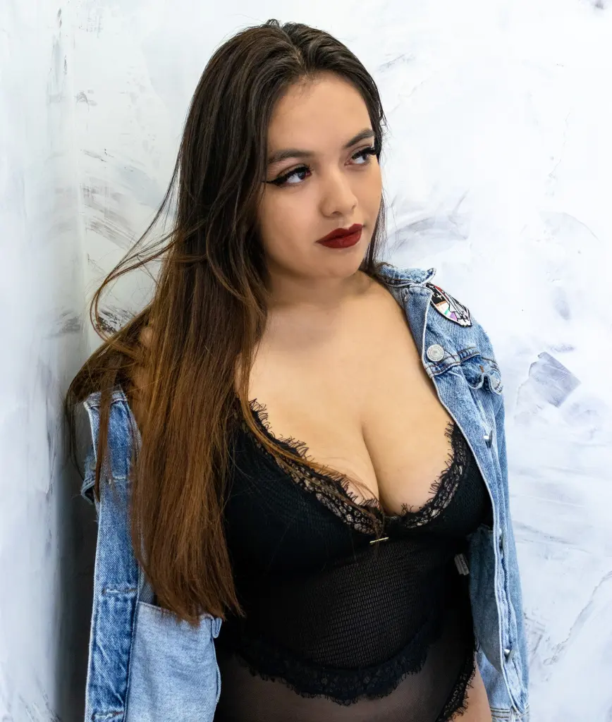 Ibizaa poses with her long dark hair down, showing her bare shoulders and chest against a plain wall.