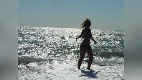 Young woman in a bikini walking through ocean waves on a sunny beach at golden hour.
