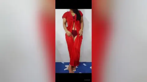 GauriKhan poses in a red open-front robe and matching red underwear, showing her stomach and pussy against a plain white wall.