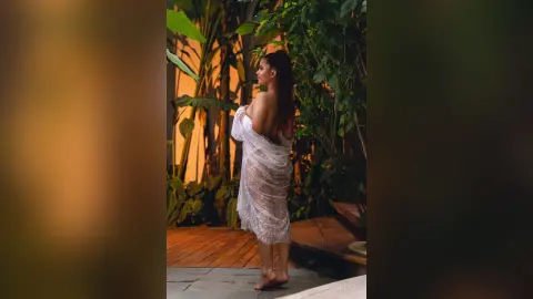 Woman in a white lace sheer cover-up standing on a wooden deck in a lush tropical garden at night.