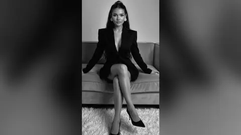 Elegant woman in a black blazer and high heels sitting on a sofa in a moody black and white studio portrait.