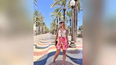 KaylaBryan stands on a wavy patterned walkway lined with palm trees, wearing a white cutout top and pink floral shorts.