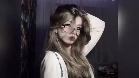 Young woman with long blonde hair wearing glasses and a white sweater in a moody, dimly lit room.