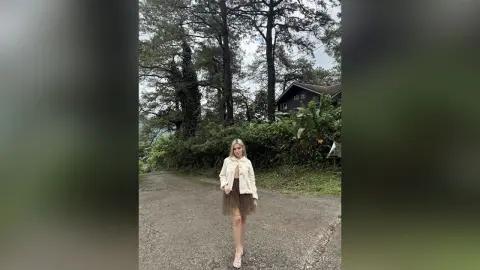 Blonde woman in a cream jacket and brown skirt standing on a forest road surrounded by lush green trees.