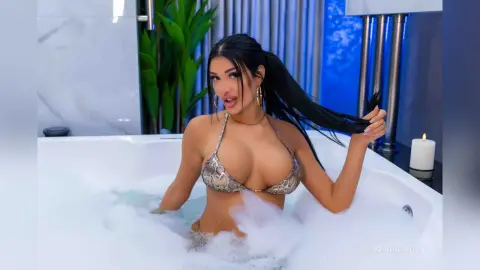 Young woman in a snakeskin bikini relaxing in a bubbly white bathtub in a modern luxury bathroom.