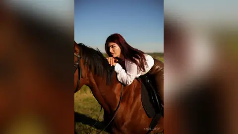 Elegant woman in a white blouse leaning on a brown horse in a sunny field during golden hour.