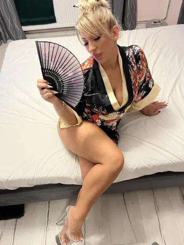 Blonde woman in a floral kimono robe sitting on a bed holding a black folding fan in a cozy bedroom setting.