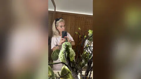 Blonde woman in a white t-shirt taking a mirror selfie next to indoor green plants in a bright room.