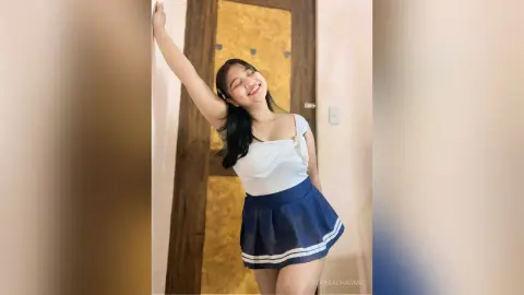 Cheerful young woman in a white tank top and navy pleated skirt posing happily in a sunlit room.