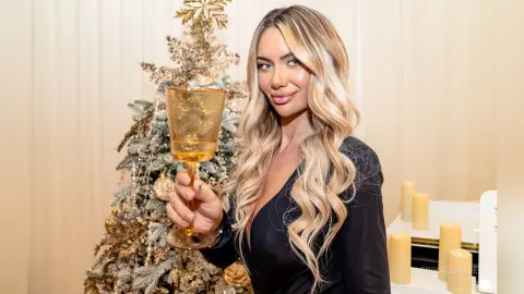 Blonde woman in a black dress holding a champagne glass near a decorated Christmas tree in a festive setting.
