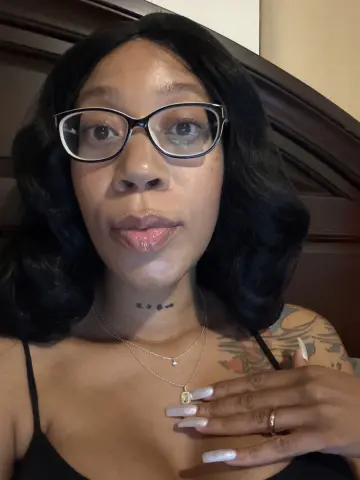 LexBright wears black glasses and a black tank top, showing off her dark wavy hair and colorful shoulder tattoo.