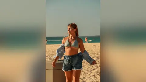 Young woman in denim shorts and bikini top relaxing on a sunny beach under a clear blue sky.
