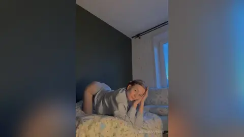 Young woman in a grey striped pajama set relaxing on a floral bed in a cozy, sunlit bedroom.
