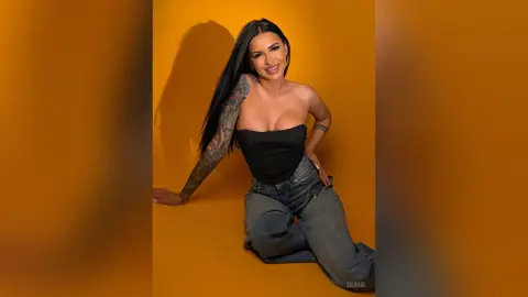 Smiling woman with long dark hair and arm tattoos wearing a black tube top and denim jeans against a yellow background.