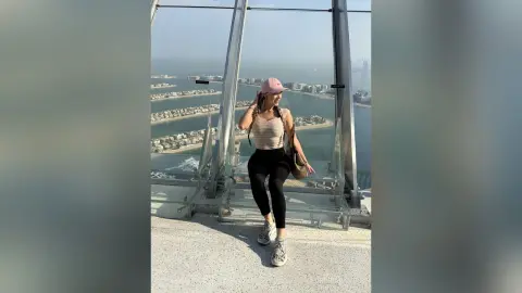 Young woman in casual tank top and leggings posing on a high-rise balcony overlooking a coastal city skyline.