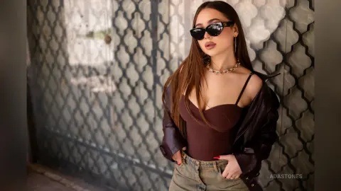 Stylish woman in a brown bodysuit and leather jacket wearing sunglasses against a patterned metal gate.