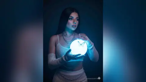 Young woman in white knit outfit holding a glowing moon orb in a moody, dark blue ethereal setting.