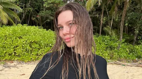 LiaMotzer poses with wet, long hair against a backdrop of tropical green bushes and palm trees.