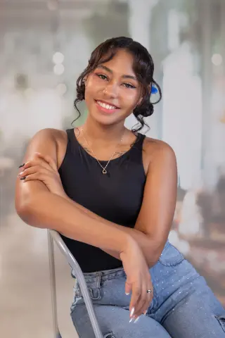 Smiling young woman in a black tank top and blue jeans sitting in a bright, modern indoor setting.