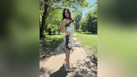 CassyTorres stands on a brick path in a park, wearing a white lace-trimmed slip dress and white high heels.