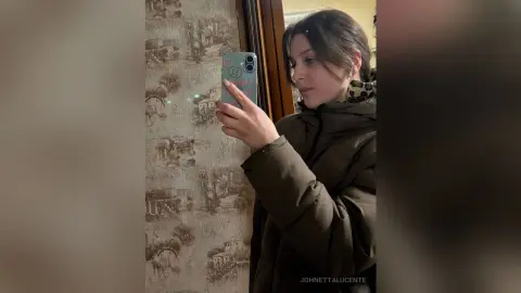 Young woman in a brown puffer jacket taking a mirror selfie in a cozy room.