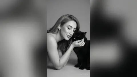 Serene black and white portrait of a woman in a strapless top cuddling a fluffy black cat in a minimalist studio.
