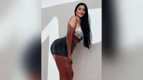 Young woman in a silver sequin bikini top and sparkly mini skirt posing against a white wall in bright sunlight.