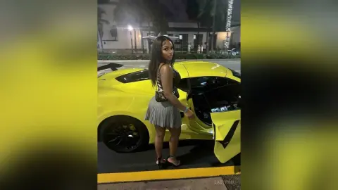 Stylish woman in a black tank top and pleated skirt standing by a bright yellow luxury sports car at night.