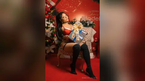 RebecaVillalobos sits on a chair wearing red lace lingerie and black thigh-high stockings while holding a wrapped gift.