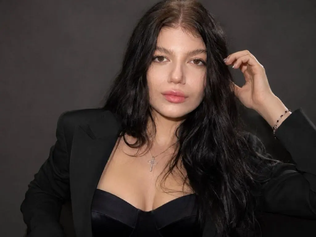 Young woman with long dark hair wearing a black blazer and corset top against a dark studio background.